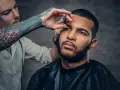 Barbero con tatuajes en el brazo recorta el **buzz cut corto** de un hombre con barba.