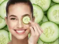 Una mujer sonríe mientras se coloca una rodaja de pepino en el ojo, rodeada de más rodajas de pepino.