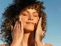 Mujer con cabello rizado aplica crema en el rostro, disfrutando del sol. El retinol en verano puede ser un aliado para una piel radiante.