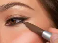 Ojo marrón aplicando delineador en la línea de agua inferior, creando un look de **reverse eyeliner** con sombra dorada en el párpado.