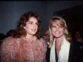 Dos mujeres con peinados icónicos de los 80, una con cabello castaño voluminoso y otra con rubio liso, posan sonrientes.