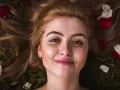 Rostro de mujer joven con pecas y cejas naturales sin depilar, rodeada de pétalos de rosa y hojas.