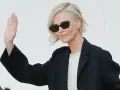 Mujer con cortes de pelo corto rubio, gafas de sol y chaqueta negra saluda.