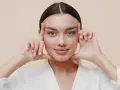 Mujer con ojos azules realizando ejercicios faciales, aplicando presión en las sienes y barbilla para tonificar.