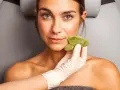 Una mano enguantada aplica una piedra de jade en la mejilla de una mujer, mostrando los beneficios del masaje facial para una piel radiante.