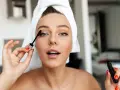 Mujer con toalla en la cabeza aplicándose máscara de pestañas como parte de su rutina maquillaje.