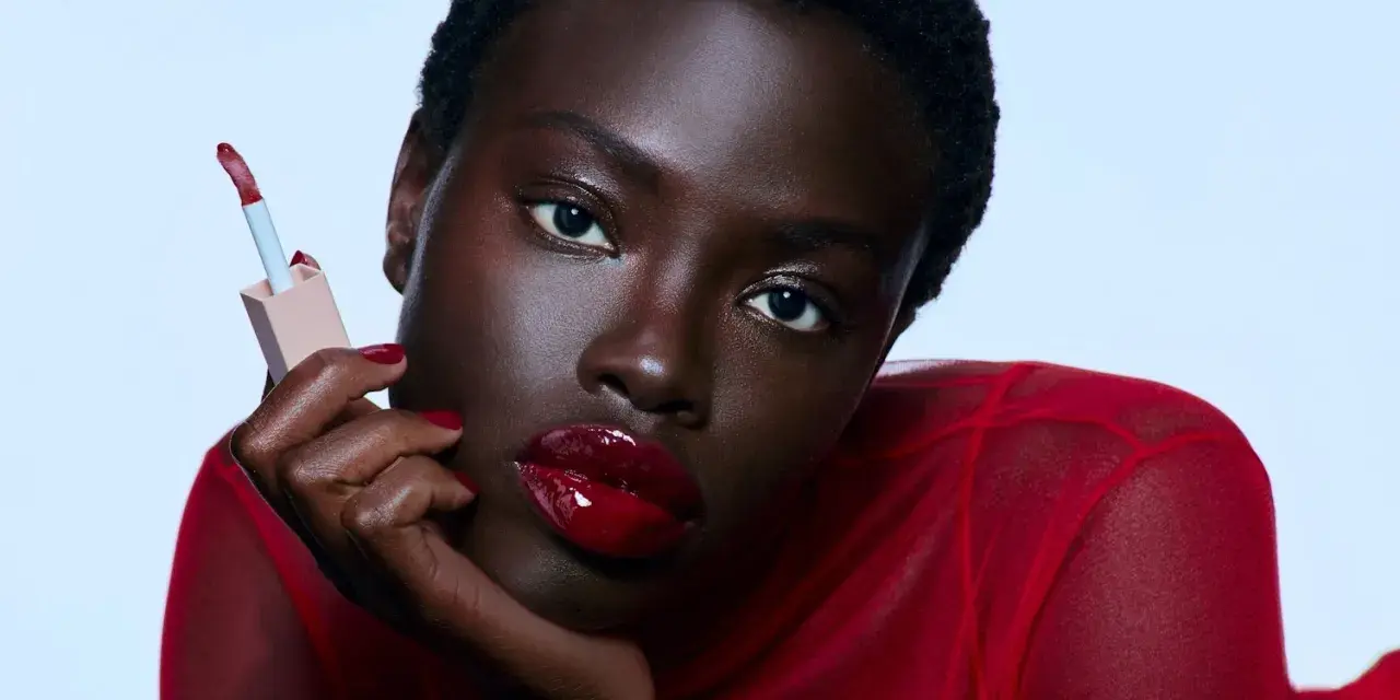 Mujer con labios rojos brillantes y un aplicador de brillo labial. Sus cejas rojas resaltan su mirada intensa.