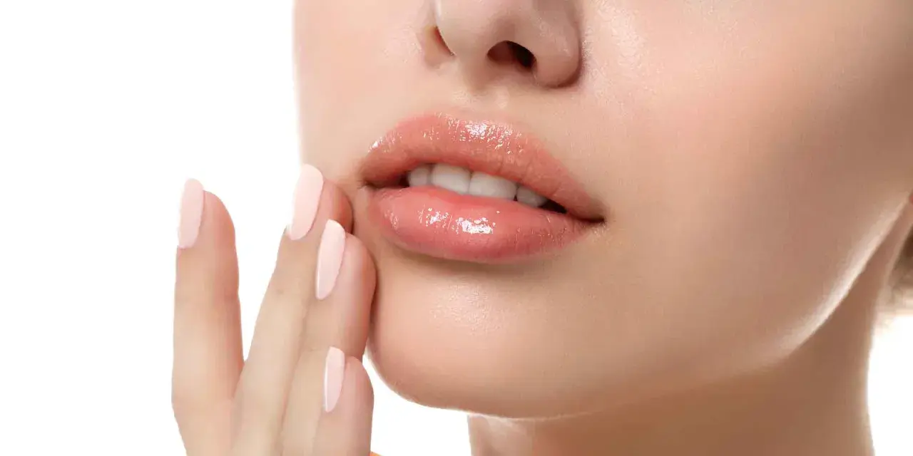 Labios rosados y brillantes, con una mano de uñas pálidas tocándolos suavemente. Un ejemplo perfecto de como hidratar los labios para un look jugoso.