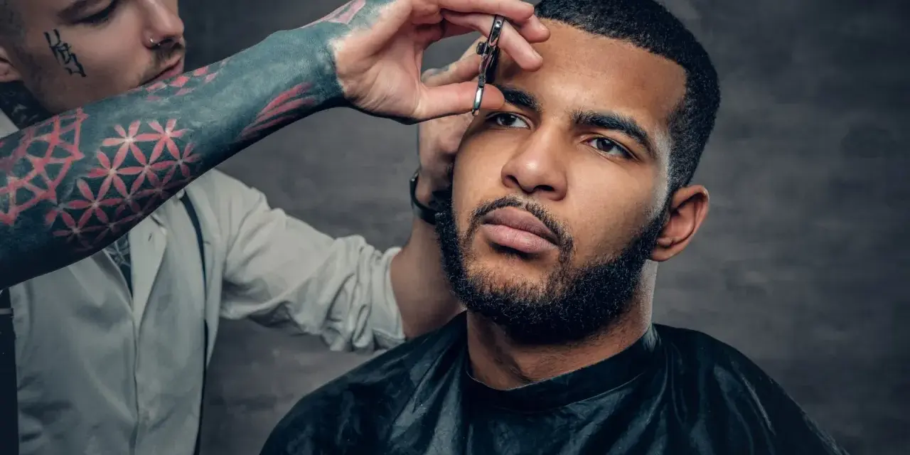 Barbero con tatuajes en el brazo recorta el **buzz cut corto** de un hombre con barba.