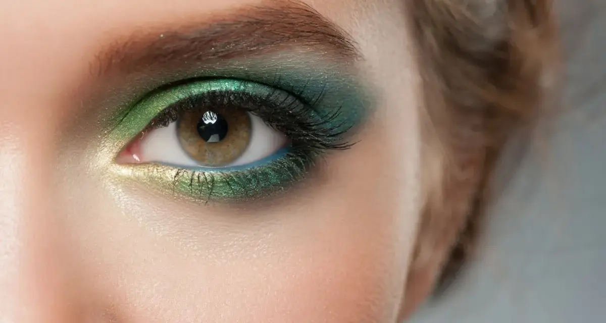 Ojo con maquillaje verde esmeralda y dorado, delineado azul y cejas verdes.