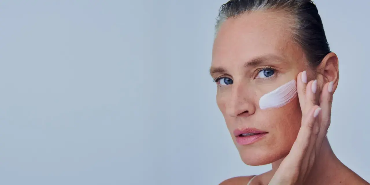 Mujer se aplica crema en la mejilla, como si fuera a quitar autobronceador. Suave toque para una piel radiante.