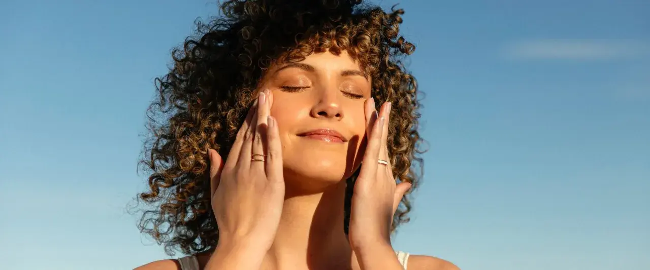 Mujer con cabello rizado aplica crema en el rostro, disfrutando del sol. El retinol en verano puede ser un aliado para una piel radiante.