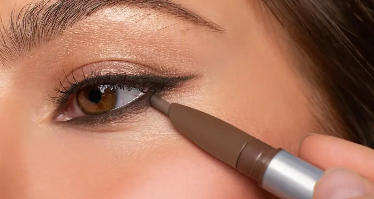 Ojo marrón aplicando delineador en la línea de agua inferior, creando un look de **reverse eyeliner** con sombra dorada en el párpado.
