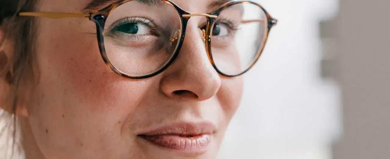 Primer plano de una mujer con gafas de montura de carey, sus ojos hundidos miran hacia un lado con una leve sonrisa.