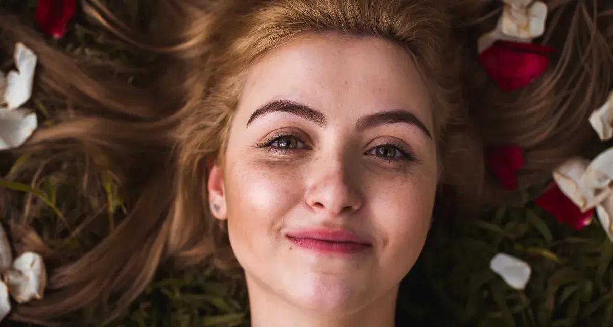 Rostro de mujer joven con pecas y cejas naturales sin depilar, rodeada de pétalos de rosa y hojas.