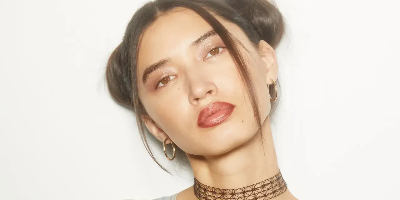Chica con peinados y maquillaje de los 90, dos moños altos, labios marrones y choker.