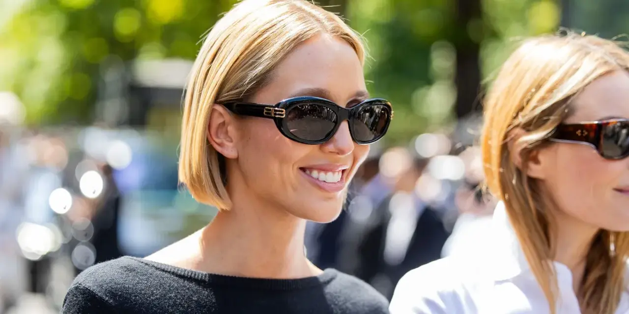 Mujer sonriente con cabello corto y cara redonda, luciendo gafas de sol negras y un suéter negro.