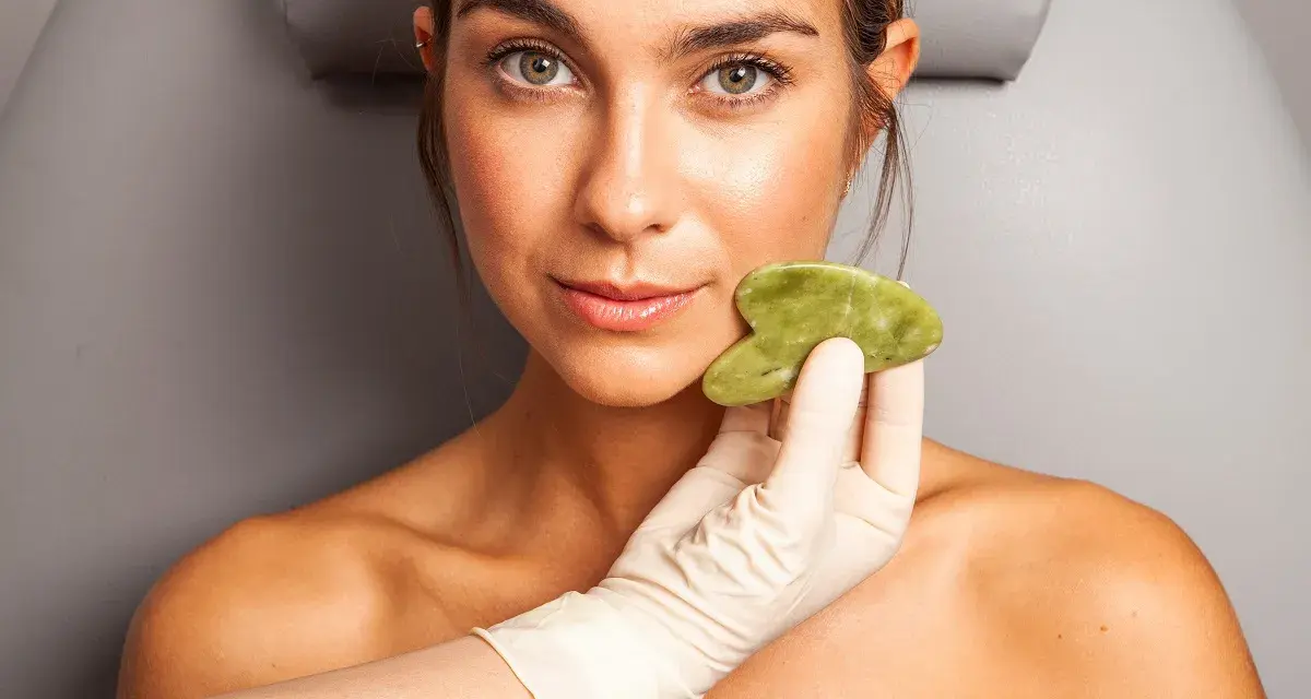 Una mano enguantada aplica una piedra de jade en la mejilla de una mujer, mostrando los beneficios del masaje facial para una piel radiante.