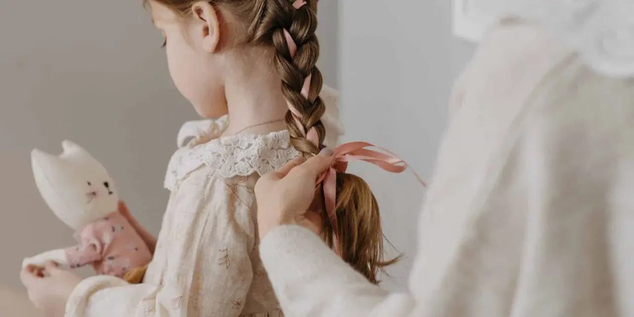 Manos terminan de trenzar el cabello de una niña, añadiendo cintas rosas. Ideas de peinados para niñas adorables.