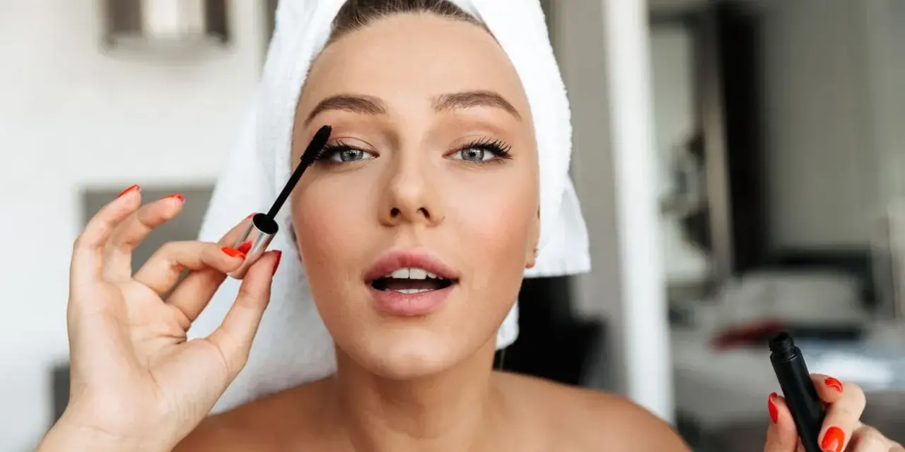 Mujer con toalla en la cabeza aplicándose máscara de pestañas como parte de su rutina maquillaje.