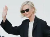 Mujer con cortes de pelo corto rubio, gafas de sol y chaqueta negra saluda.