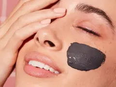 Una mujer sonríe con una mascarilla facial de arcilla negra en la mejilla. Después de una mascarilla facial, hay que ponerse crema para hidratar la piel.