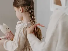 Manos terminan de trenzar el cabello de una niña, añadiendo cintas rosas. Ideas de peinados para niñas adorables.