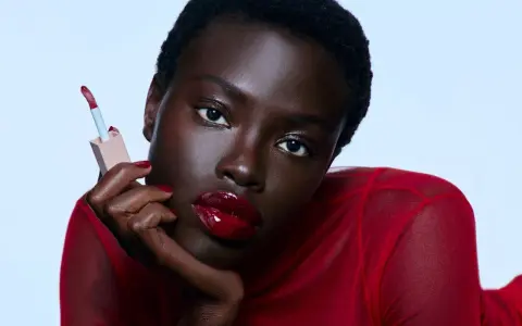 Mujer con labios rojos brillantes y un aplicador de brillo labial. Sus cejas rojas resaltan su mirada intensa.