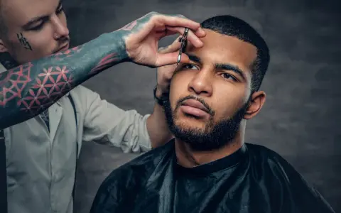 Barbero con tatuajes en el brazo recorta el **buzz cut corto** de un hombre con barba.