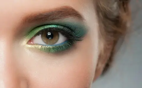 Ojo con maquillaje verde esmeralda y dorado, delineado azul y cejas verdes.