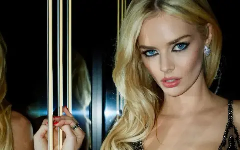 Maquillaje ojos verdes boda: rubia con ojos azules intensos, labios rosados y vestido negro brillante.