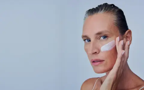Mujer se aplica crema en la mejilla, como si fuera a quitar autobronceador. Suave toque para una piel radiante.