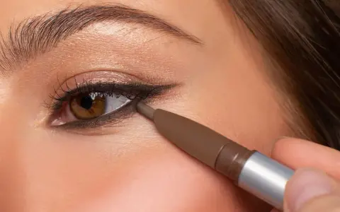 Ojo marrón aplicando delineador en la línea de agua inferior, creando un look de **reverse eyeliner** con sombra dorada en el párpado.
