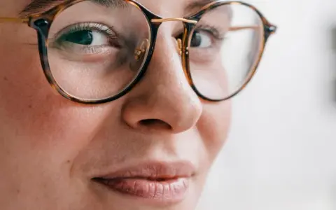 Primer plano de una mujer con gafas de montura de carey, sus ojos hundidos miran hacia un lado con una leve sonrisa.