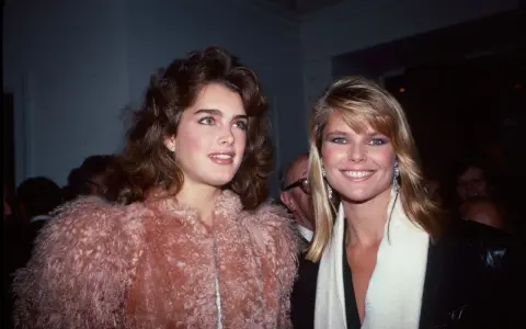 Dos mujeres con peinados icónicos de los 80, una con cabello castaño voluminoso y otra con rubio liso, posan sonrientes.