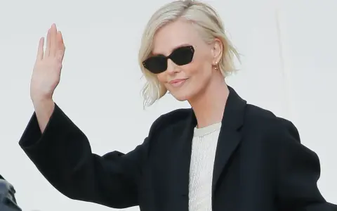Mujer con cortes de pelo corto rubio, gafas de sol y chaqueta negra saluda.