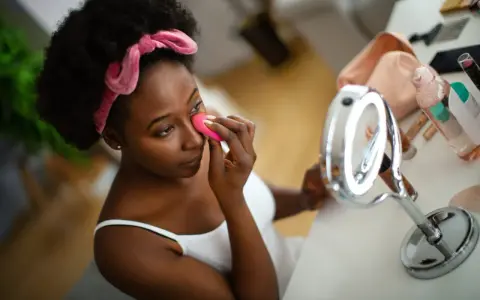 Una mujer se aplica base de maquillaje con esponja frente a un espejo.