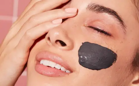 Una mujer sonríe con una mascarilla facial de arcilla negra en la mejilla. Después de una mascarilla facial, hay que ponerse crema para hidratar la piel.