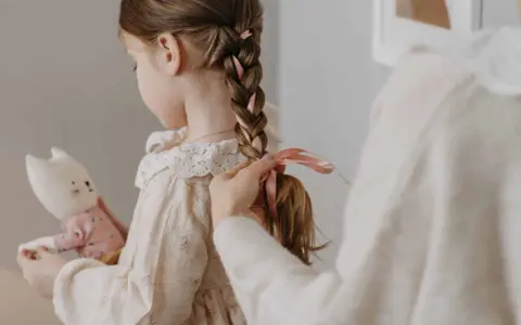 Manos terminan de trenzar el cabello de una niña, añadiendo cintas rosas. Ideas de peinados para niñas adorables.