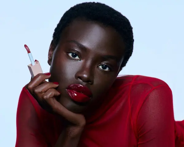 Mujer con labios rojos brillantes y un aplicador de brillo labial. Sus cejas rojas resaltan su mirada intensa.