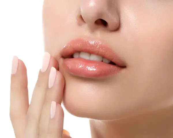 Labios rosados y brillantes, con una mano de uñas pálidas tocándolos suavemente. Un ejemplo perfecto de como hidratar los labios para un look jugoso.