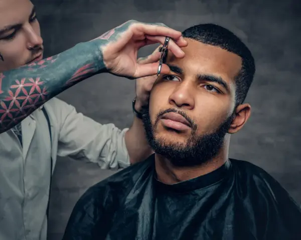 Barbero con tatuajes en el brazo recorta el **buzz cut corto** de un hombre con barba.
