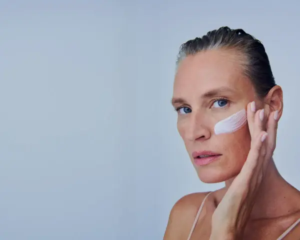 Mujer se aplica crema en la mejilla, como si fuera a quitar autobronceador. Suave toque para una piel radiante.