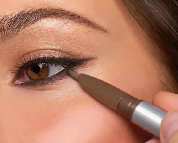 Ojo marrón aplicando delineador en la línea de agua inferior, creando un look de **reverse eyeliner** con sombra dorada en el párpado.