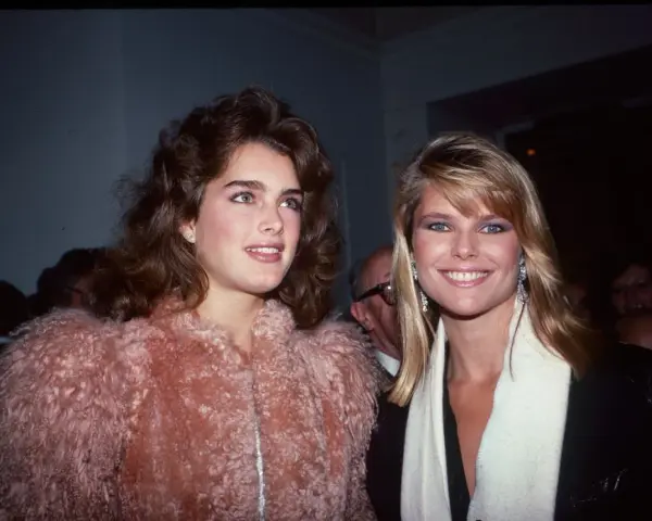 Dos mujeres con peinados icónicos de los 80, una con cabello castaño voluminoso y otra con rubio liso, posan sonrientes.