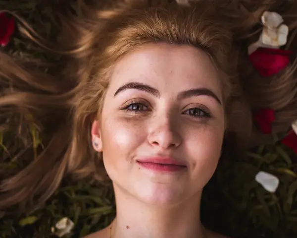 Rostro de mujer joven con pecas y cejas naturales sin depilar, rodeada de pétalos de rosa y hojas.