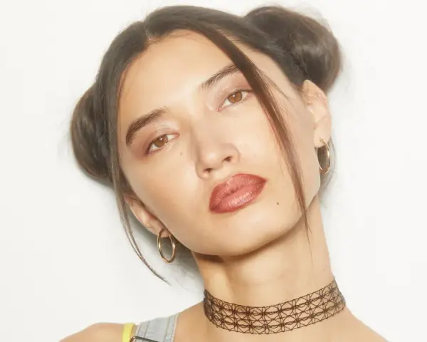 Chica con peinados y maquillaje de los 90, dos moños altos, labios marrones y choker.