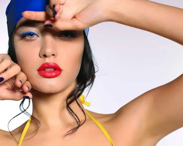 Mujer con gorro azul y traje de baño amarillo, labios rojos y delineado azul vibrante. Un look de maquillaje para ir a la playa.