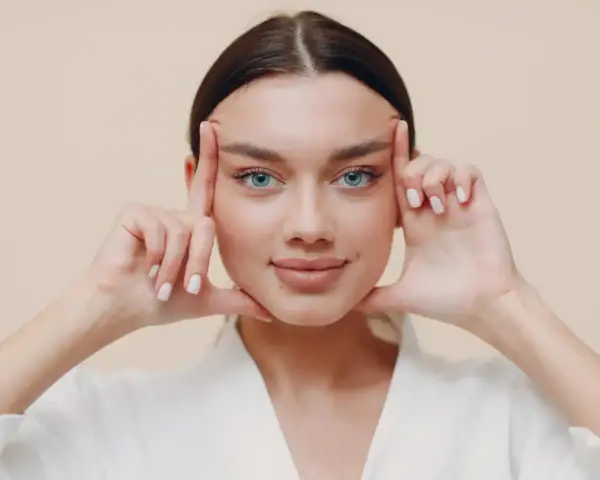 Mujer con ojos azules realizando ejercicios faciales, aplicando presión en las sienes y barbilla para tonificar.