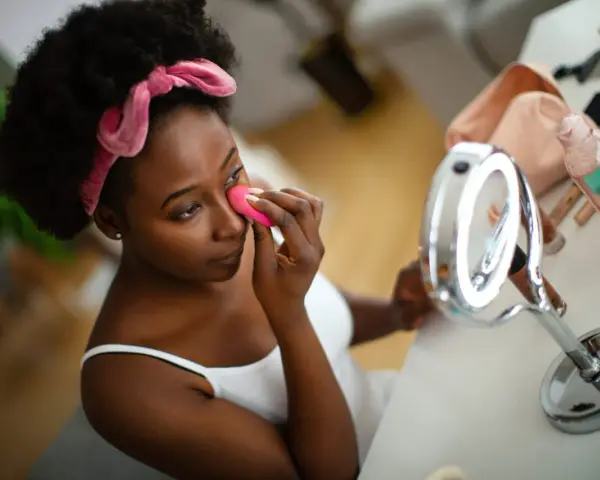 Una mujer se aplica base de maquillaje con esponja frente a un espejo.